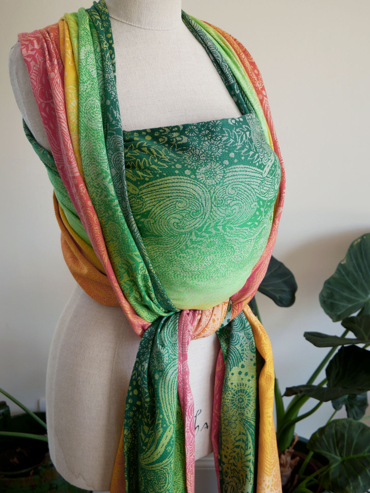 Mannequin wearing a colourful Marina Starburst Baby Wrap with a green patterned top against a white background