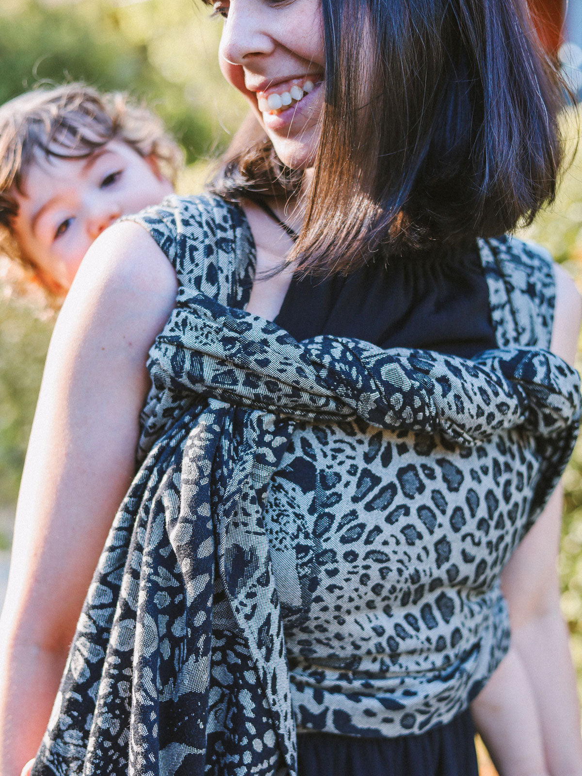 Woman wearing a leopard print Prowl Savannah Baby Wrap with a child in the background