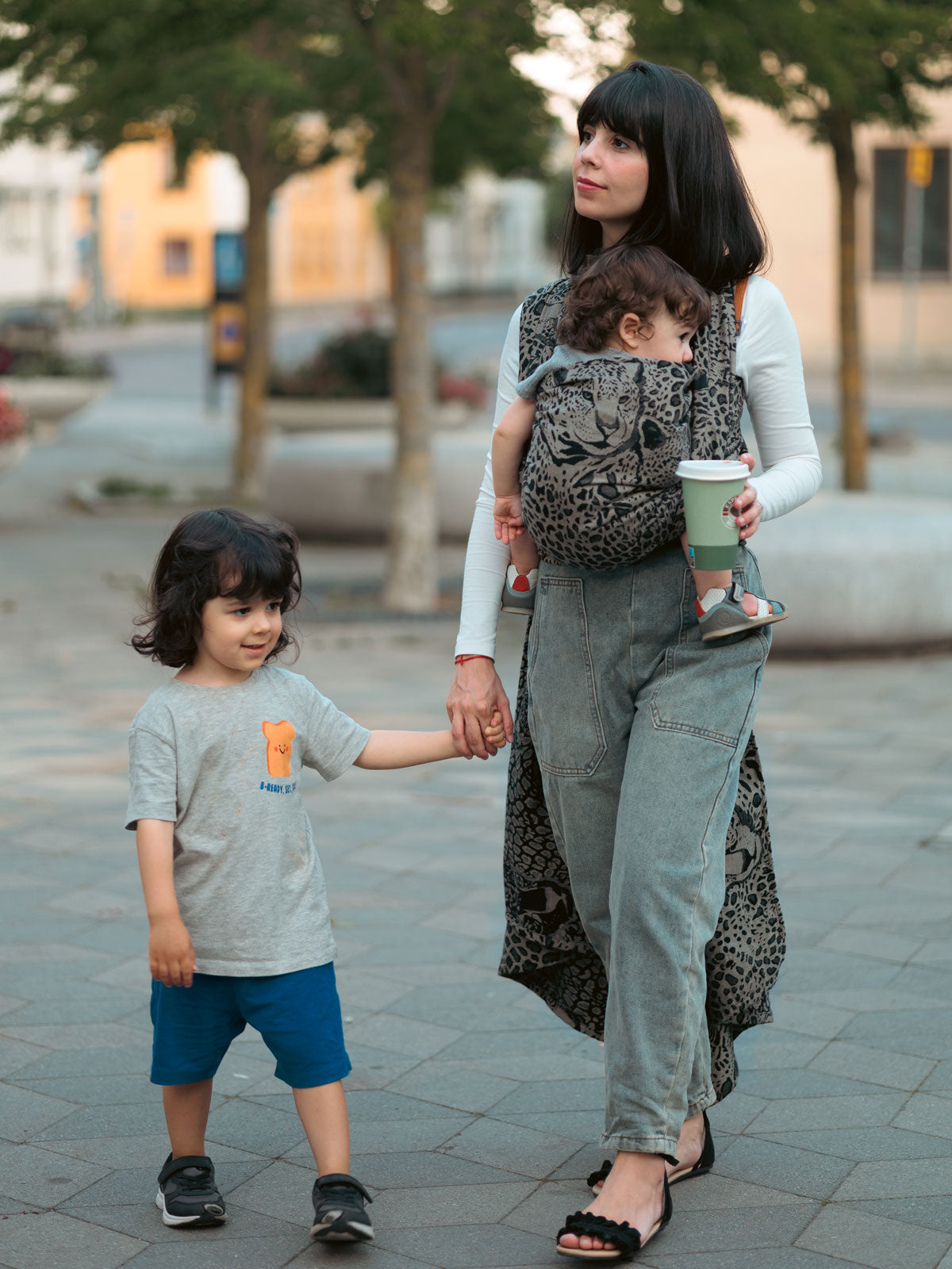Woman walking with a child in Prowl Savannah Baby Wrap and another child holding hands on a sidewalk.