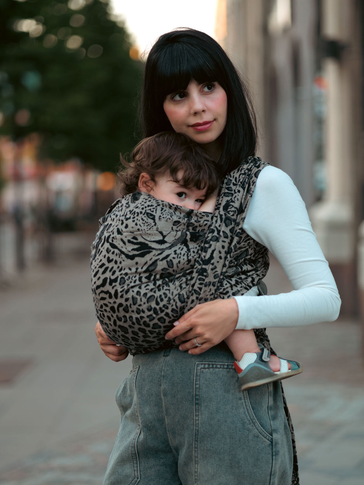 Woman carrying a child in a leopard print sling on a city street.