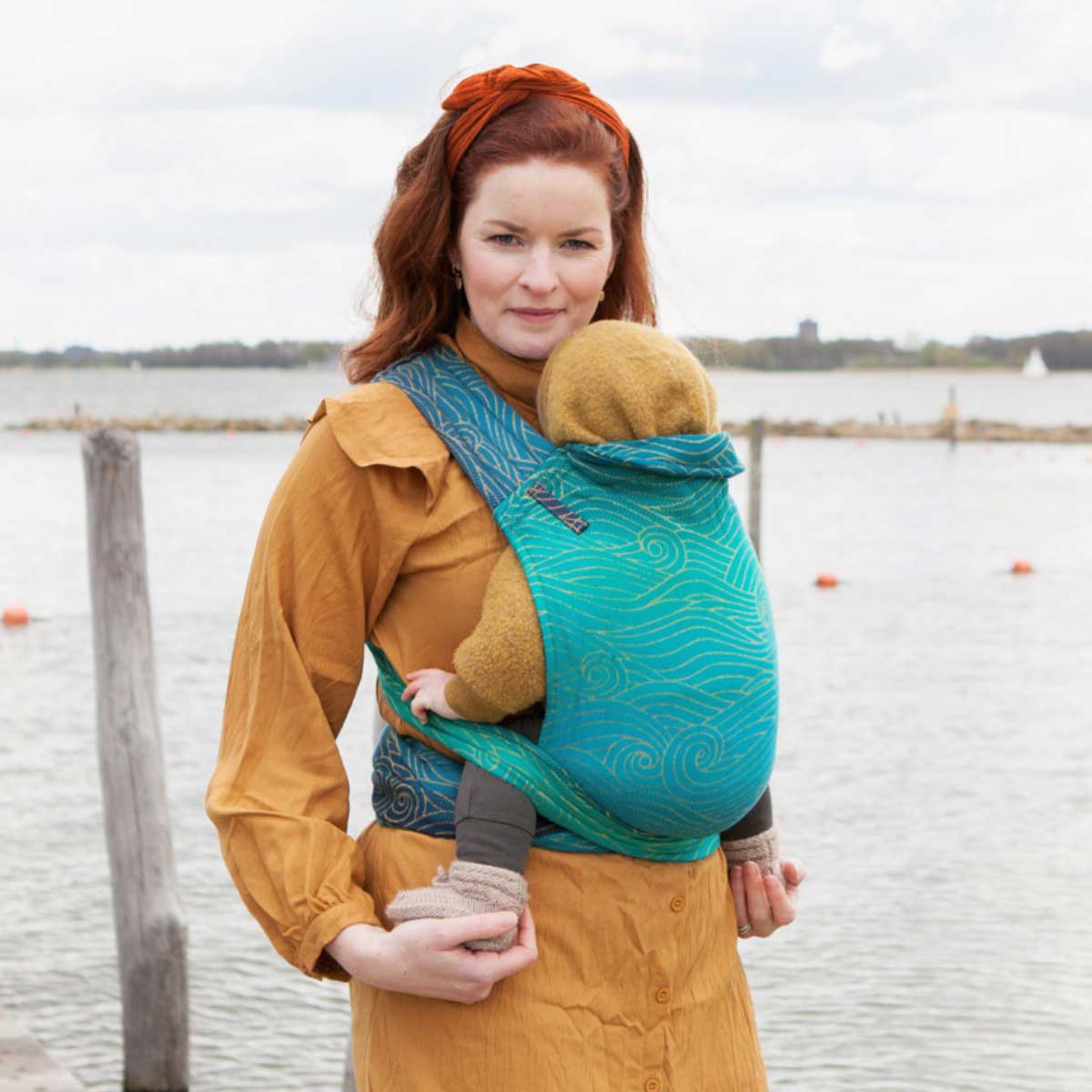 Woman carrying a child in a teal baby carrier by a waterfront