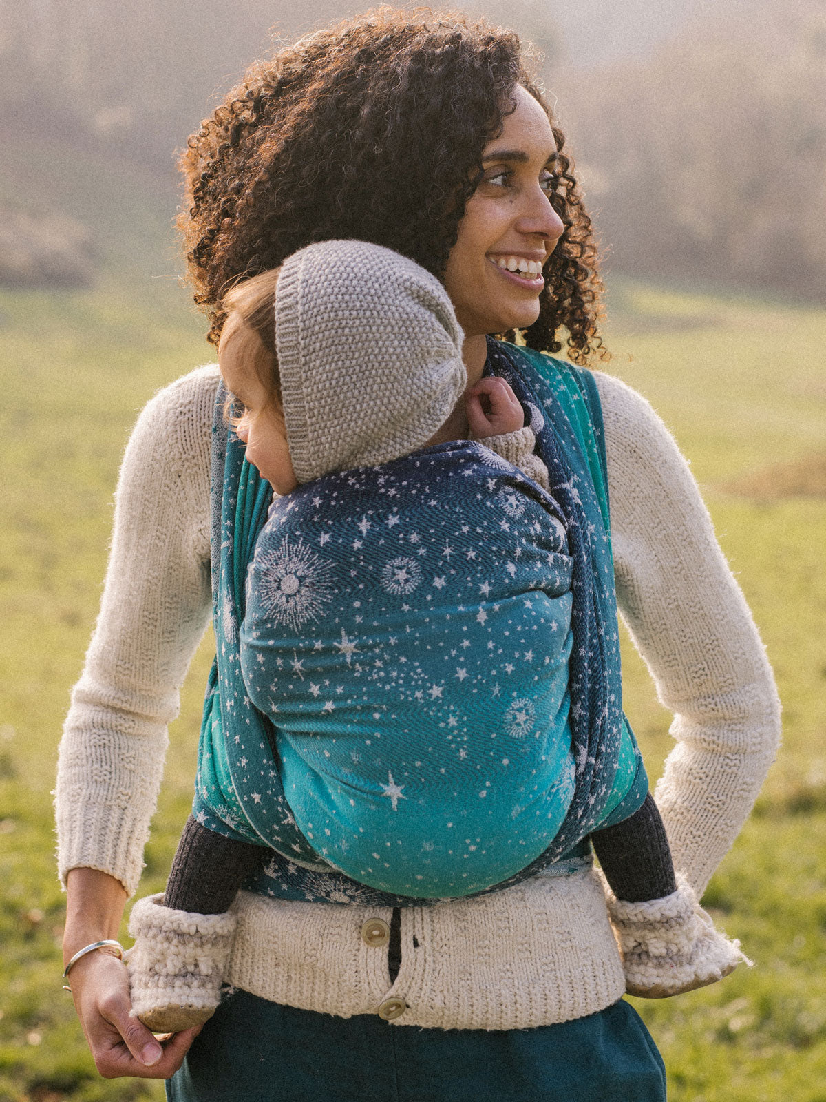 A person is outdoors holding a baby in a blue baby wrap with white stars and constellations design.