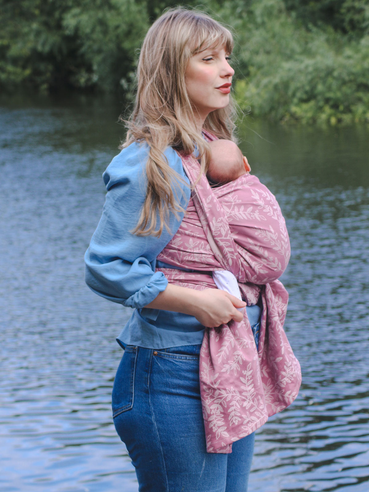 Woman carrying a baby in a pink sling by a body of water.